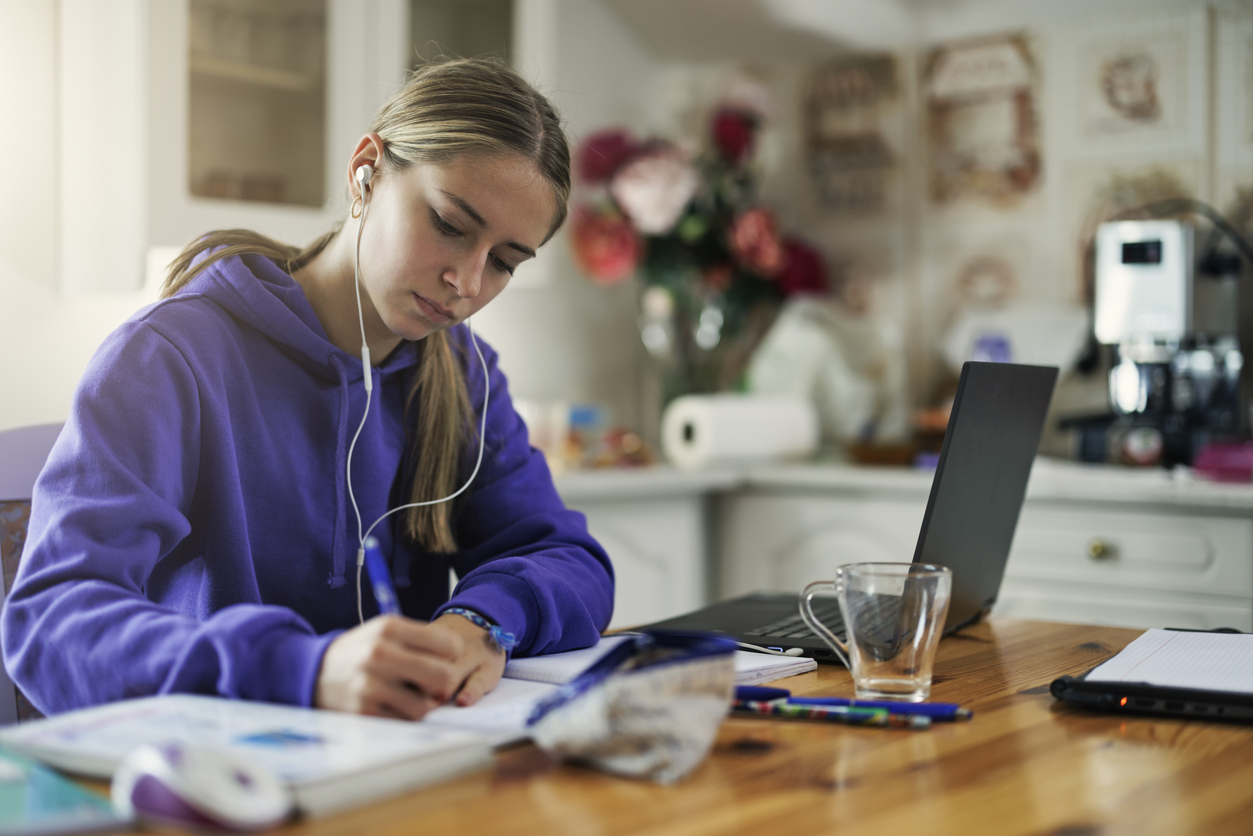A teen student in headphones writing notes while studying at a kitchen table